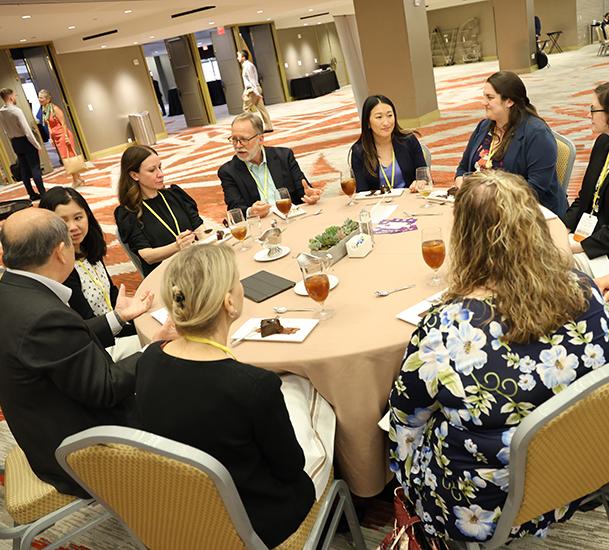 variety of individuals seated around a round table enjoying food and beverages and socializing